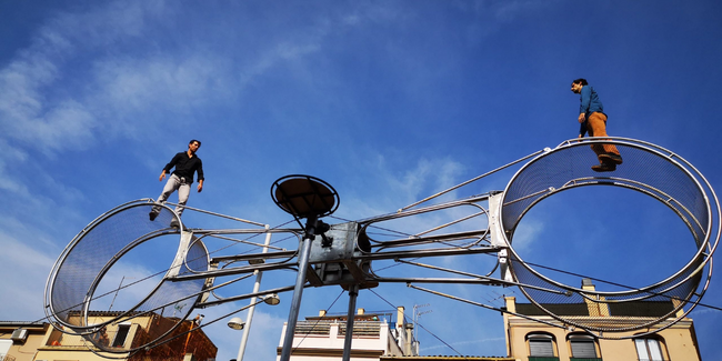 2 artistes de cirque réalisent un numéro d'équilibriste sur 2 roues énormes rattachées à un système métallique.